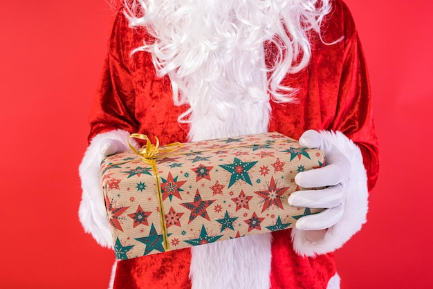 Detail van de handen van een man verkleed als kerstman met handschoenen die een geschenk vasthoudt op een rode achtergrond Kerstviering geschenken consumentisme en geluk concept