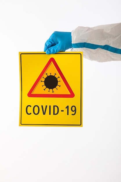 Detail van een arm van een medisch verpleegkundige die een PBM en latexhandschoenen draagt met een geel bord met een COVID-19-gevaarsymbool met de tekst: 'COVID-19'. Coronavirus, pandemie en gezondheidsconcept.