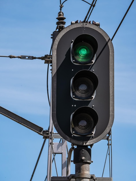 Detail view of a high railway light signal traffic light on a pole giving the upper indication of green color on freeway and the other two lamps off on a blue sky background