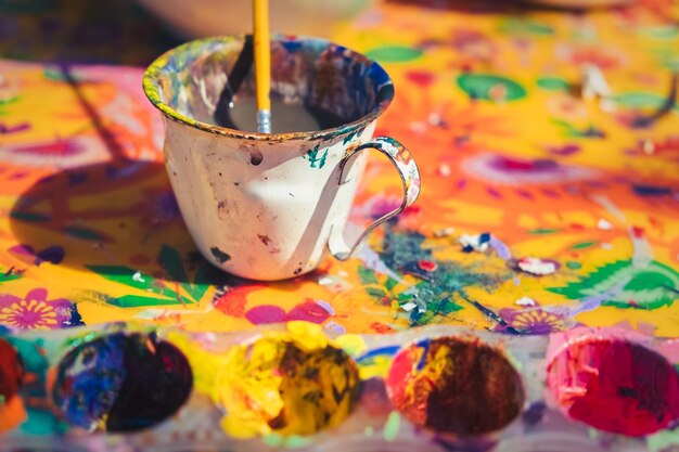 Detail view of messy painting colours and painter's cup with paintbrush on colorful canvas