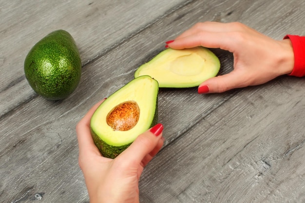 Detail on young woman hands with red nails, holding avocado cut in half over gray wood desk, with other whole one in background.