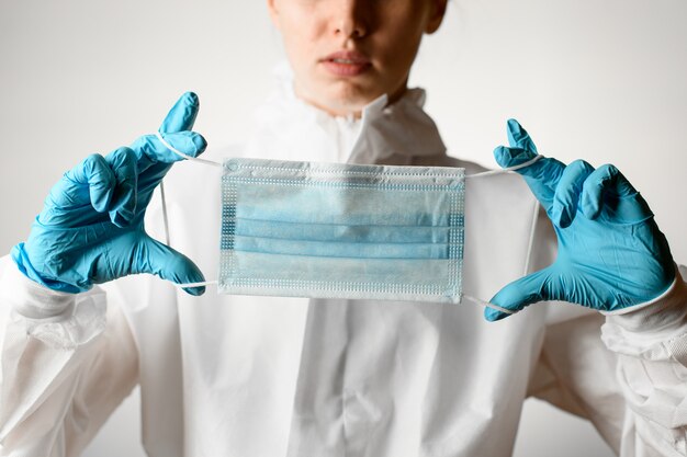 Detailopname. Vrouw in wit beschermend pak en handschoenen houdt medische masker in haar handen.