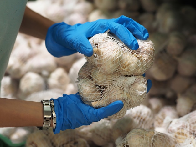 Detailopname. Vrouwelijke handen in blauwe handschoenen nemen een knoflook in de supermarkt tijdens pandemisch coronavirus COVID-19.