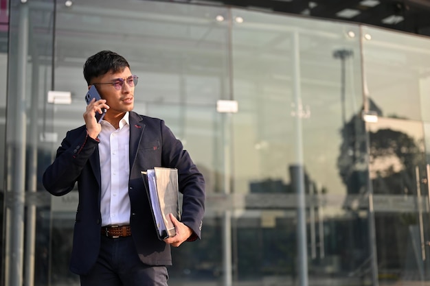 Determined Asian businessman talking on the phone while walking out of the company building