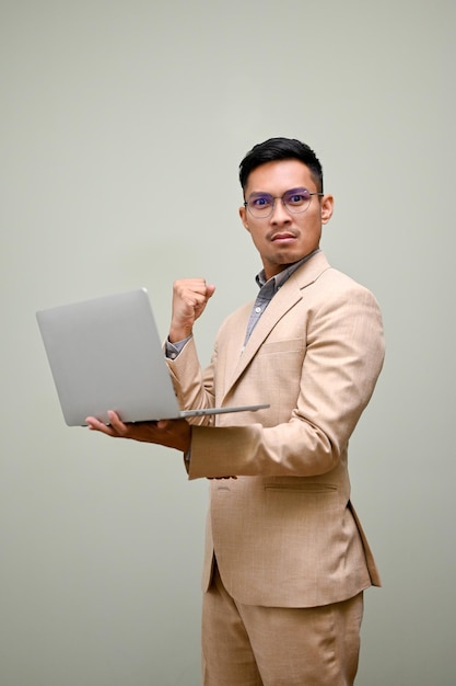 Determined Asian businessman with laptop showing clenched fist standing against green background