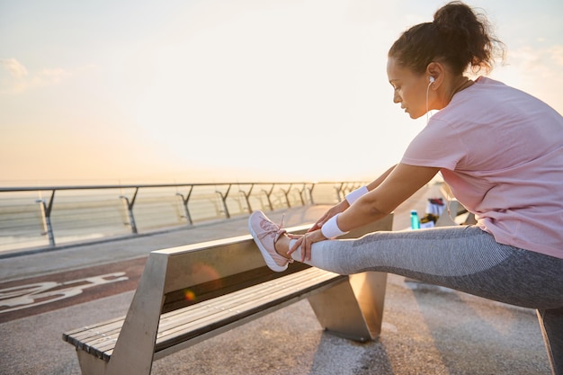 Determined female athlete, sportswoman stretching legs before morning jog. Sportswoman exercising outdoor, standing on an urban environment and flexing her legs muscles on a wooden bench at sunrise