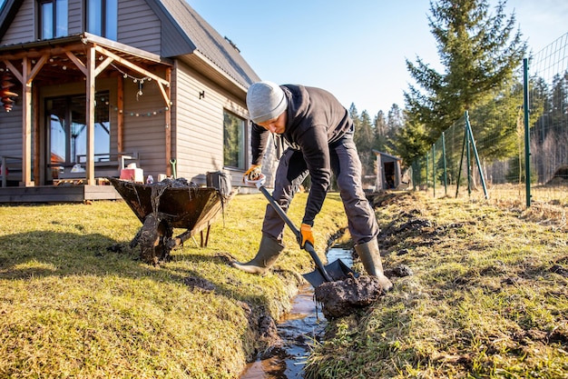 Решительный мужчина лопатует грязь из траншеи на своем заднем дворе в солнечный день с тележкой и сельским домом на заднем плане