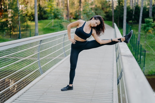 Determined sportswoman stretches legs on bridge, wears active wear, demonstrates good flexibility, poses outdoor against forest wall. Woman runner prepares for jogging, has summer workout