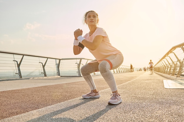 Determined sporty woman exercising outdoor at sunrise, doing\
squats with elastic resistance band. fitness, sport, endurance,\
body weight training and active lifestyle concept