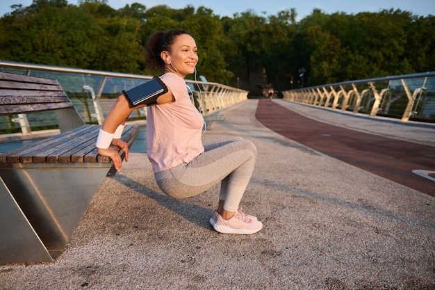 Determined young sportswoman enjoying her morning workout\
outdoor, doing exercises on triceps, flexing her arms on a wooden\
bench while practicing a body weight training on the city bridge at\
sunrise