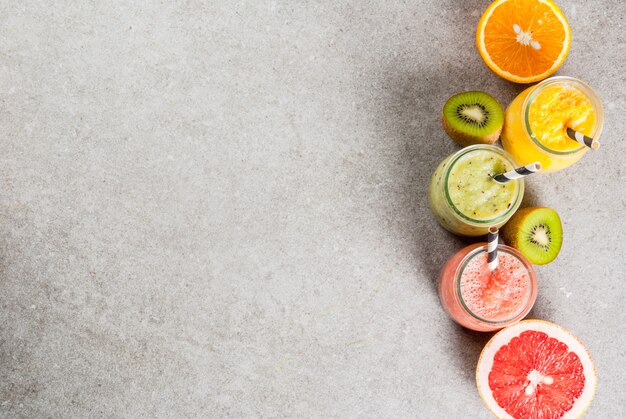 Detox biologische dieetdranken, zelfgemaakte tropische smoothies - kiwi, sinaasappel, grapefruit, in geportioneerde potten, met ingrediënten, op een grijze stenen tafel. copyspace bovenaanzicht