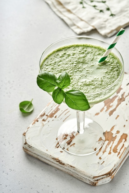 Detox green vegetable juice or smoothie garnished with leaf of fresh basil in coctail glass on light gray slate, stone or concrete background. Top view with copy space.