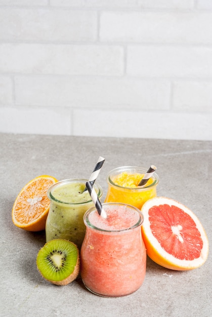 Photo detox organic diet drinks, homemade tropical smoothies - kiwi, orange, grapefruit, in portioned jars, with ingredients, on a gray stone table.  copyspace