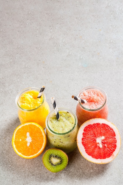 Detox organic diet drinks, homemade tropical smoothies - kiwi, orange, grapefruit, in portioned jars, with ingredients, on a gray stone table.  copyspace