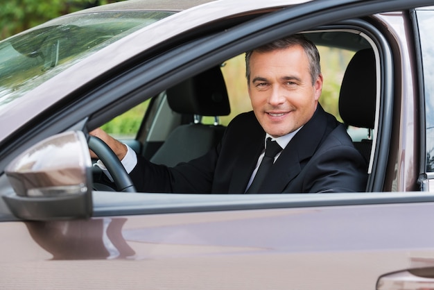 Deze auto is gewoon perfect! Vrolijke volwassen man in formalwear die naar de auto kijkt en glimlacht terwijl hij in de auto zit