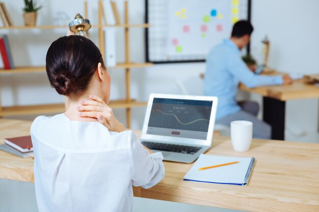 Do diagram analysis. Serious businesswoman sitting at her workplace and touching neck while taking decision