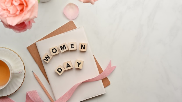 Dice with word "Women day" on notebooks decorated with pink flowers, ribbon and copy space on marble table