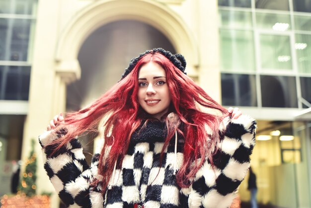 Dichtbij portret van een glimlachend tienermeisje met rood haar in warme kleren die buiten staan en in de camera kijken en met eigen rood haar spelen.