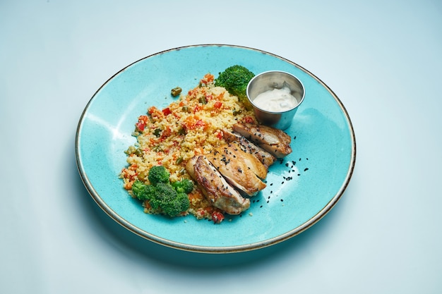 Dieet en gezond voedsel gebakken kippendijen met couscous, broccoli in een blauwe keramische plaat geïsoleerd op grijs oppervlak. Detailopname