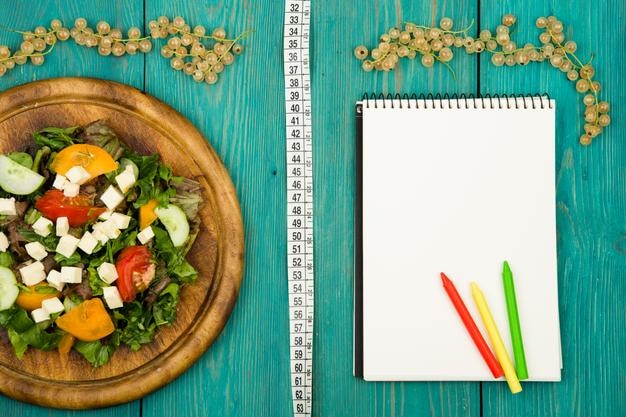 Foto dieetplansalade van meetlint met verse groenten en blanco notitieboekje op een witte houten tafel