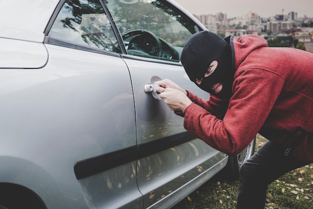 Dief die binnen een autoraam kijkt klaar om iets te stelen. Crimineel in zwarte bivakmuts en hoodie opent iemands voertuig met loper. Auto inbraak door onbekende man met verborgen gezicht.