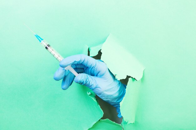 Dien een blauwe medische handschoen met een spuit in. Door een gat in groen papier. geneeskunde, vaccinatie, gezondheid. Minimalisme,.