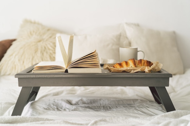 Dienblad met croissant, kopje koffie en boek op wit schoon bed.