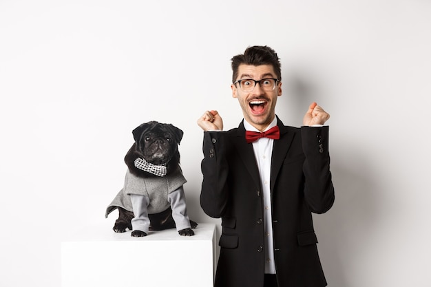 Dieren, feest en feest concept. Gelukkig jonge man in pak en puppy in huisdier cosume staande over wit, hondeneigenaar verheugend en triomfantelijk.