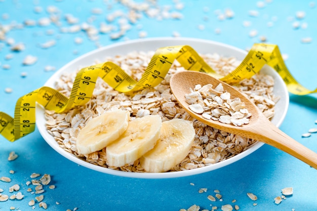 Diet concept. Oatmeal bowl and spoon with fresh ripe banana and yellow measuring tape on blue background