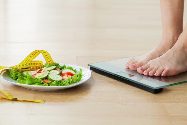 Foto concetto di dieta, la donna misura il peso su bilance elettroniche e insalata dietetica con nastro di misurazione giallo.