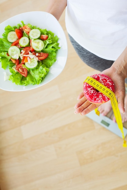 Concetto di dieta, la donna misura il peso su una bilancia elettronica mentre tiene la ciambella calorica.