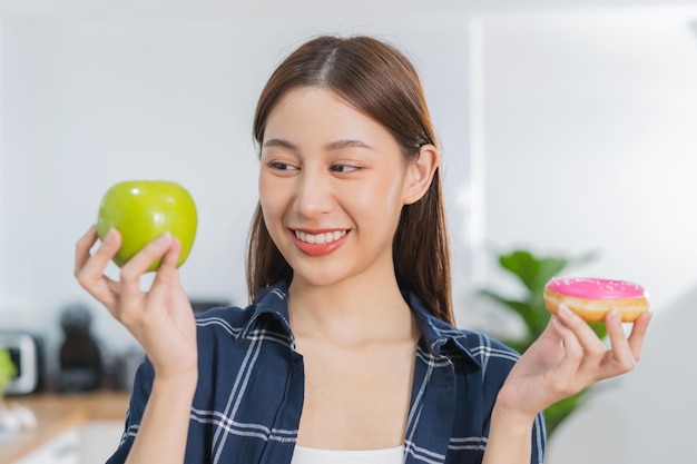 Diet Dieting asian young woman or girl smile confused choose choice green apple or pink sweet donut at home eat food for good healthy health when hungry Close up female weight loss person