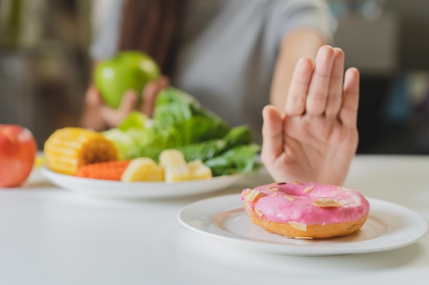 다이어트, 다이어트를 하는 젊은 아시아 여성이나 소녀는 손을 내밀고, 달콤한 도넛을 거부하고, 녹색 사과, 샐러드 야채를 선택하고, 배고플 때 건강하고 건강을 위해 음식을 먹습니다. 여성 체중 감량 사람을 닫습니다.