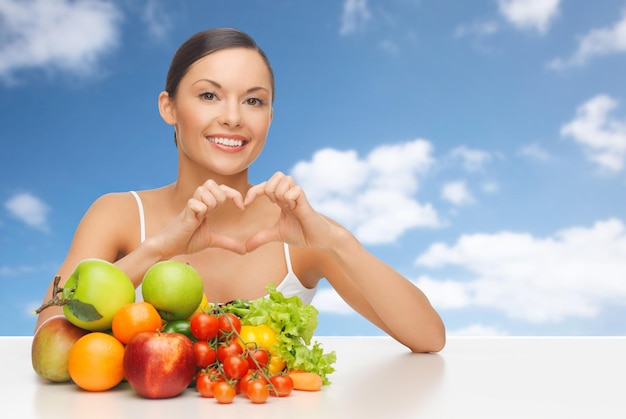 diet, healthy eating, food and people concept - happy woman with lot of fruits and vegetables showing heart over blue sky and clouds background