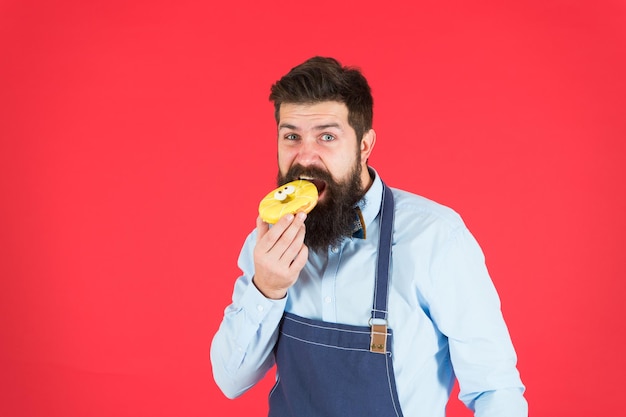 Diet and healthy food. Unhealthy eating. Baker eat doughnut. Chef man in cafe. Calorie. Feel hunger. Bearded baker. Bearded man in apron. baker hold donut. Funny hipster. Professional baker.