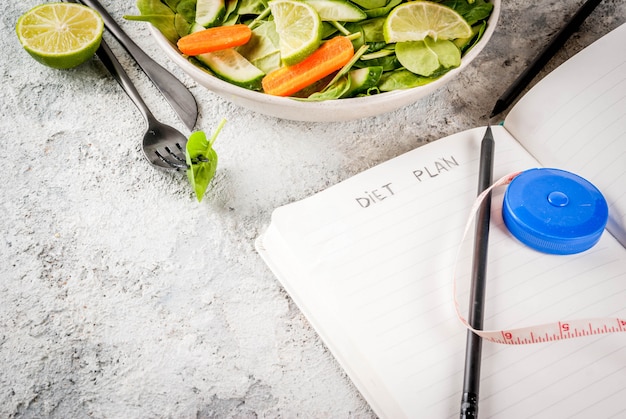 Diet plan weight lose concept, fresh vegetable salad with fork, knife, note pad,  grey stone table copy space