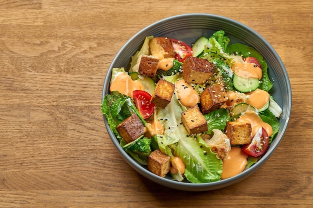 Diet salad with fried tofu, yellow sauce and vegetables in a bowl on a wooden background