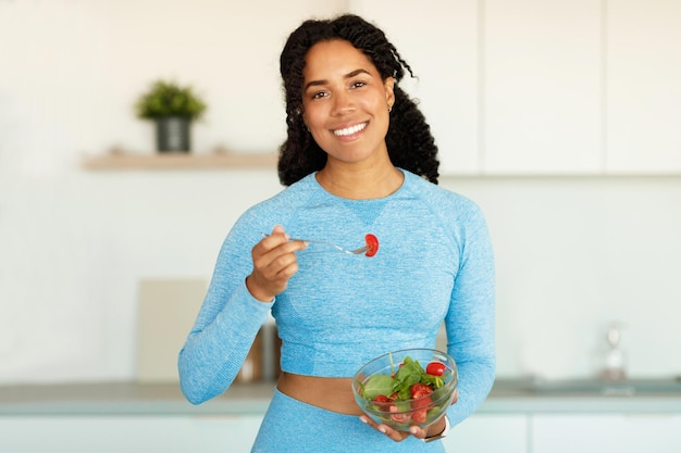 Photo diet and weight loss concept happy fit black woman in sportswear eating fresh vegetable salad in kitchen interior