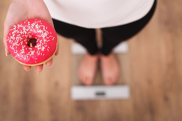 Diet, woman measuring body weight on weighing scale holding donut