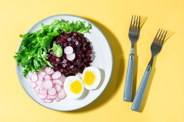 A dietary dish made from vegetables. Beet tartare, radish, frieze salad and boiled egg on a plate and a fork on a yellow table. Directly above