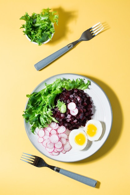 A dietary dish made from vegetables. Beet tartare, radish, frieze salad and boiled egg on a plate and a fork on a yellow table. Vertical and top view