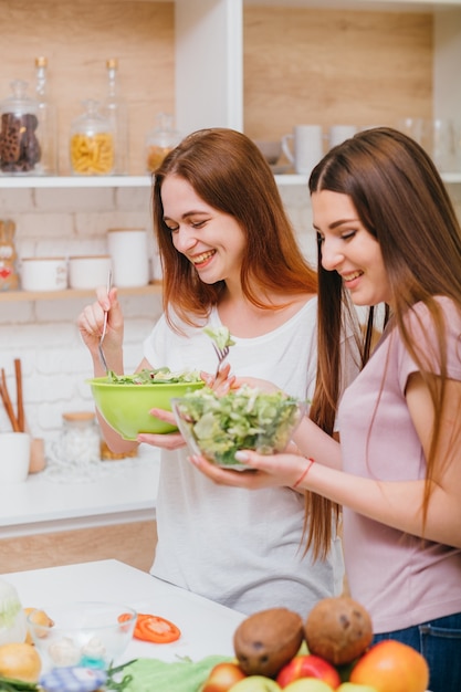 Dieting together. Young female cooking fun. Two attractive women with salad bowls. Smiling having good time.