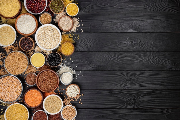 Different cereals, grains, seeds, groats, legumes and beans in bowls, top view of raw porridge collection on black wooden background with copy space