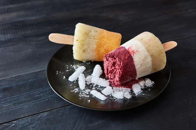 Different coffee fruit ice cream on a stick are presented in a black plate with slices of ice on a dark wooden background with space for text Healthy sweet dessert