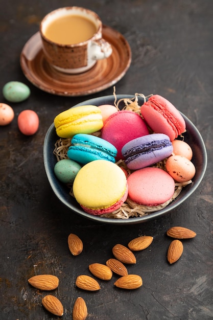 Different colors macaroons and chocolate eggs in ceramic bowl cup of coffee on black concrete background side view close up