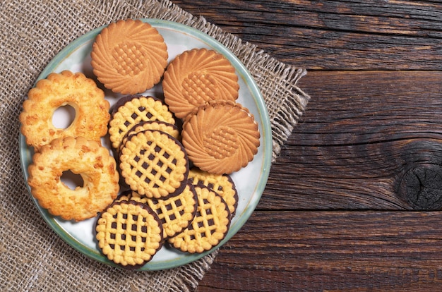 Different cookies in plate on rustic wooden table with burlap, top view