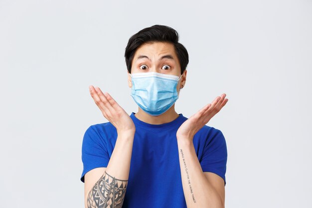 Different emotions, lifestyle and leisure during coronavirus, covid-19 concept. Surprised and astonished asian man in medical mask react to interesting gossip, raise hands near face stare camera.