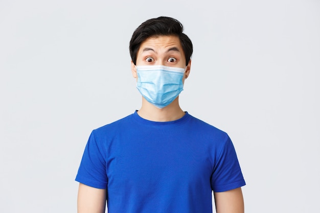 Different emotions, lifestyle and leisure during coronavirus, covid-19 concept. Surprised expression of asian guy in medical mask. Close-up guy raise eyebrows in surprise, listen news