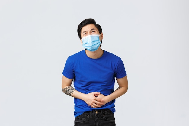 Different emotions, social distancing, self-quarantine on coronavirus and lifestyle concept. Asian man in medical mask feeling sick, have stomachache, catch food poison or flu