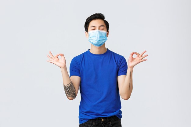 Different emotions, social distancing, self-quarantine on covid-19 and lifestyle concept. Asian guy keep relaxed and patient, meditating in medical mask, show zen signs and close eyes, do yoga